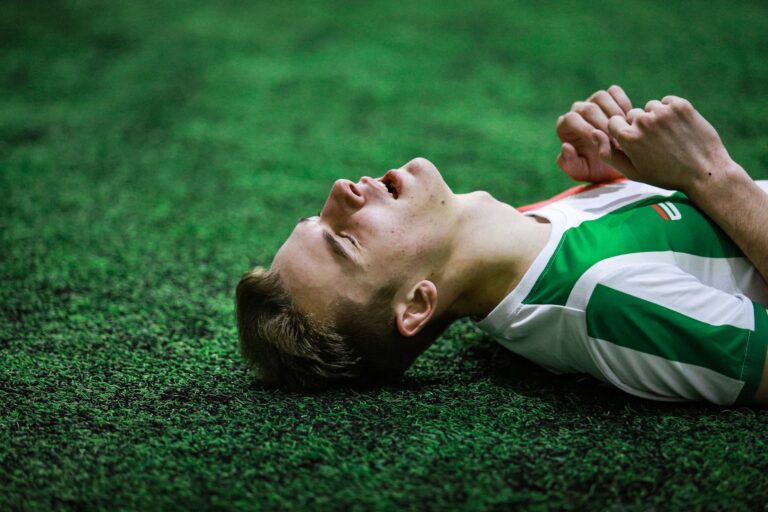 Young male soccer player in green jersey lying on grass, eyes closed, exhausted.