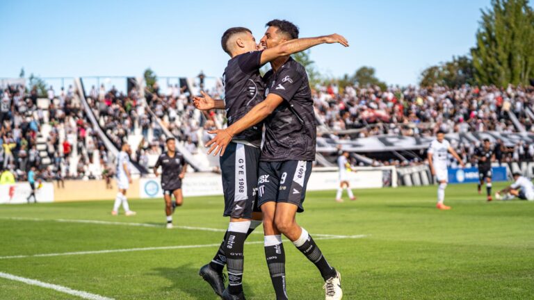 Two soccer players in black jerseys celebrate a goal on a sunny day at a packed stadium.