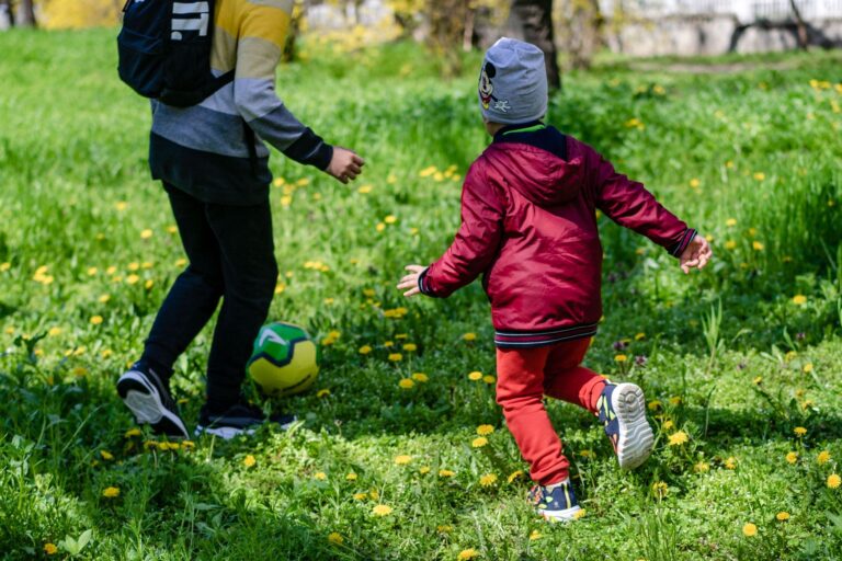 Two children enjoy a game of soccer in a sunny, grassy park area surrounded by dandelions.
