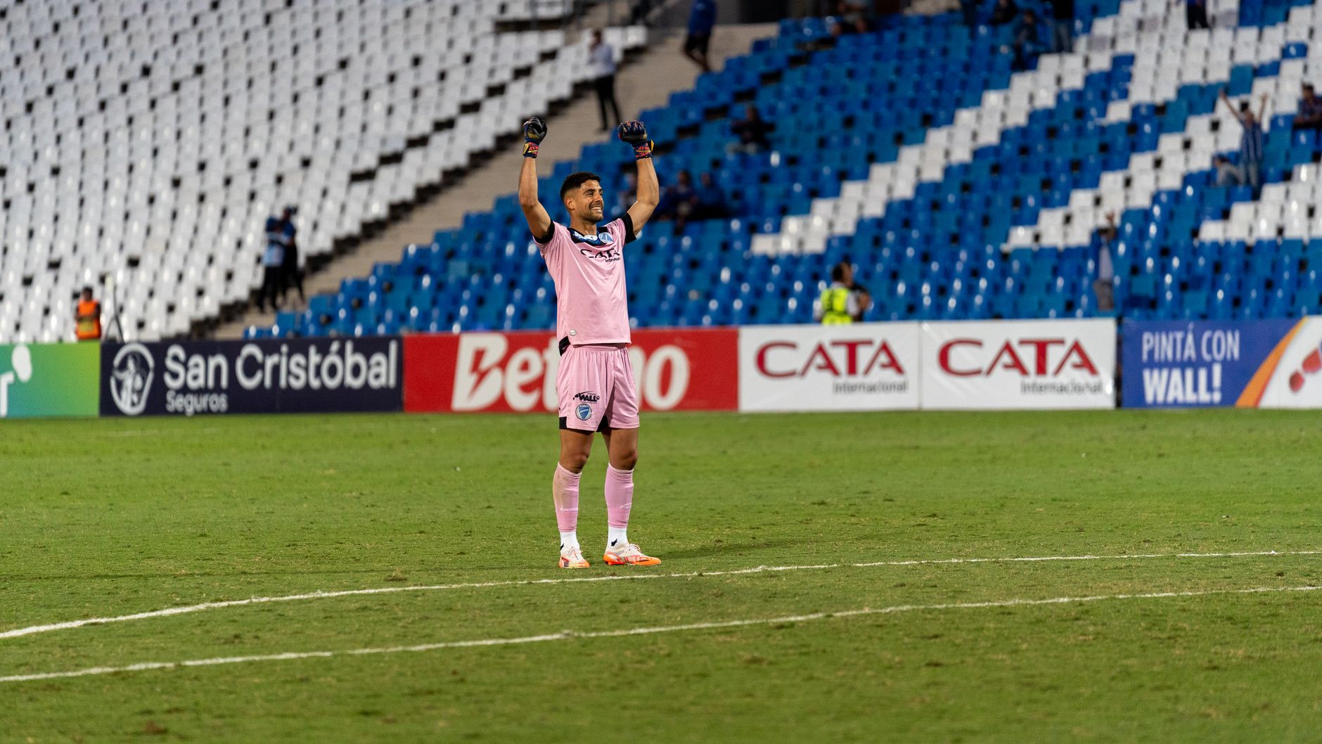Soccer goalkeeper in pink jersey celebrating victory on the field with raised arms.