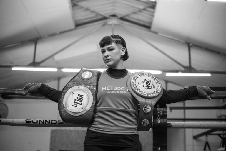 A female boxer confidently poses with championship belts in a boxing ring, showcasing strength and achievement.