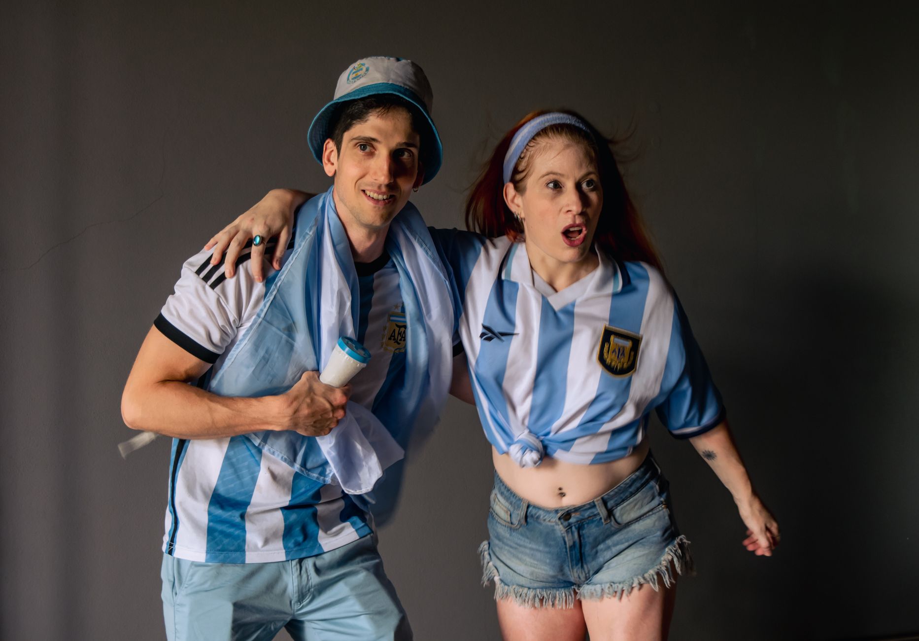 Two cheerful Argentina soccer fans celebrate wearing team jerseys indoors.
