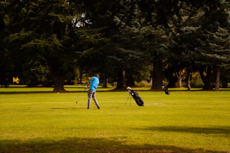 A golfer takes a swing on a lush course, surrounded by trees on a bright day.