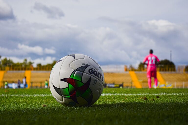Close-up of a soccer ball on green grass field with a blurred player in pink jersey in the background during a match.