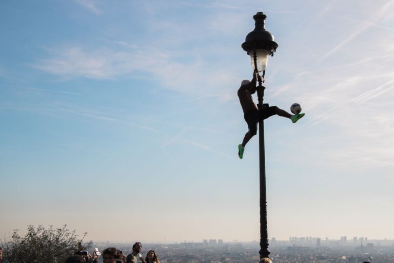 Silhouette of a man performing football tricks on a lamppost in Paris, with cityscape in the background.