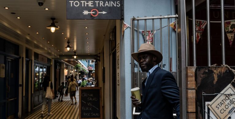 A stylish man in a suit and fedora stands next to 'To the Trains' sign in a busy London passage.