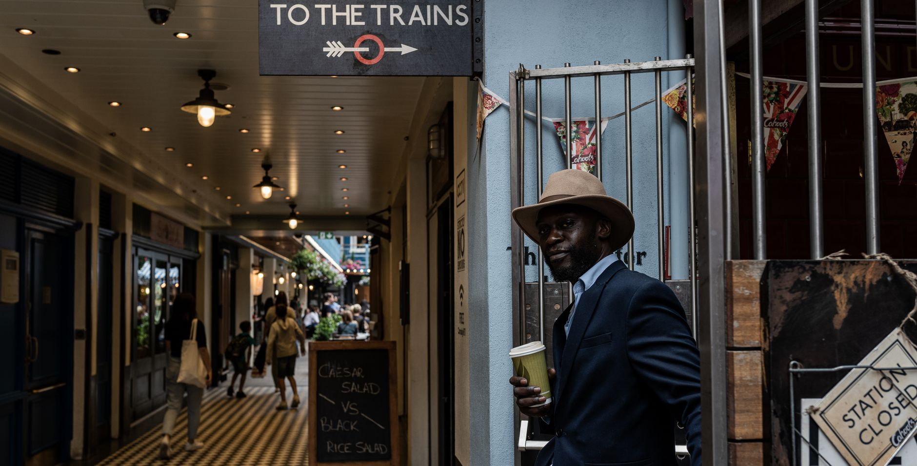A stylish man in a suit and fedora stands next to 'To the Trains' sign in a busy London passage.