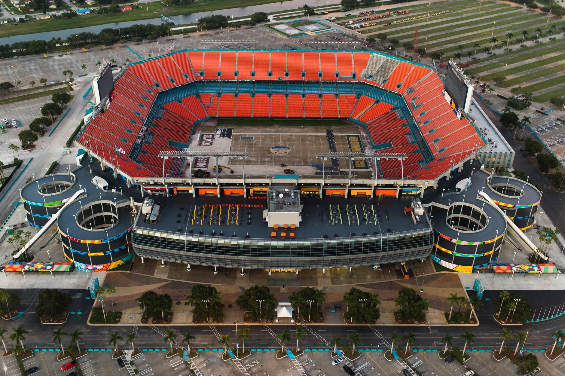 High-angle aerial shot of an empty sports stadium with orange seats and surrounding parking areas.