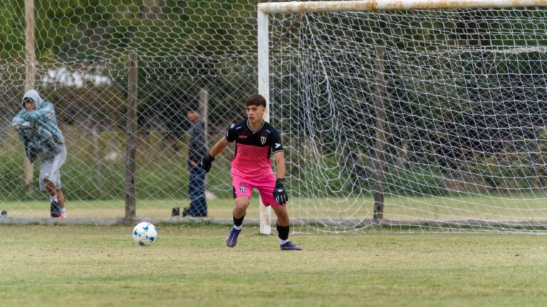 A young male goalkeeper in pink uniform guards the net during a soccer game outdoors on a grassy field.