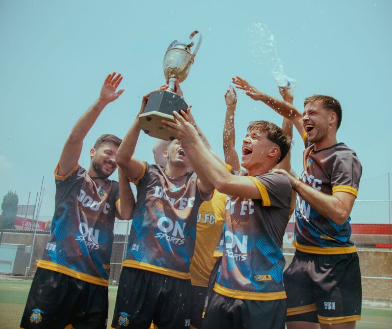 A joyous soccer team celebrates with a trophy outdoors under a clear sky.