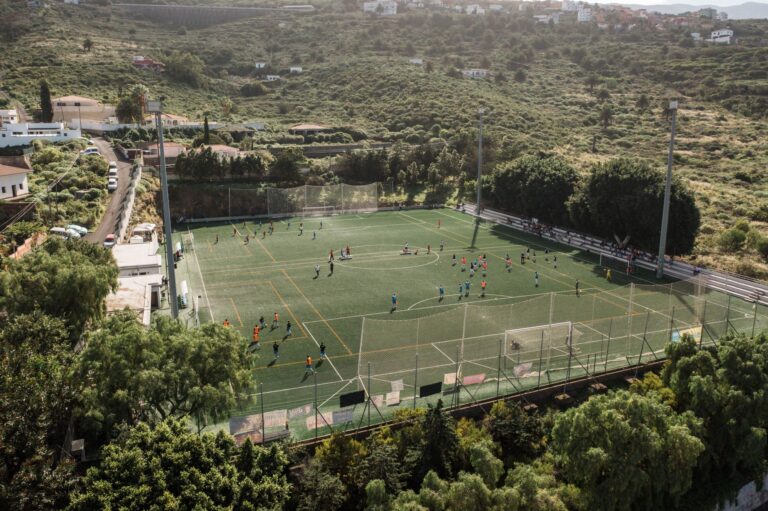 Aerial shot of soccer match on a rural field in El Sauzal, Spain, surrounded by lush greenery.