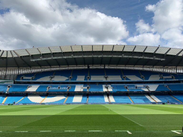 Etihad Stadium in Manchester with empty blue seats under a cloudy sky.