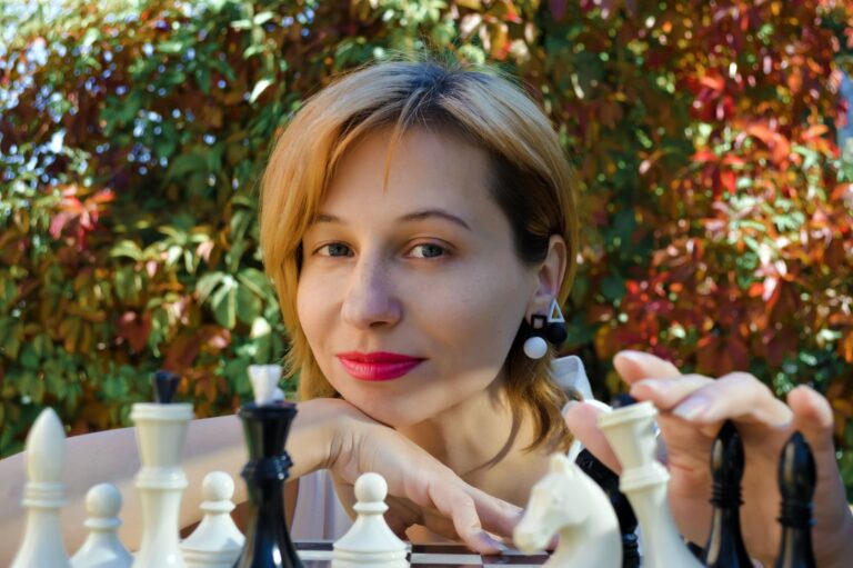 Content female player looking at camera while sitting at chess board during game on street with colorful foliage on trees