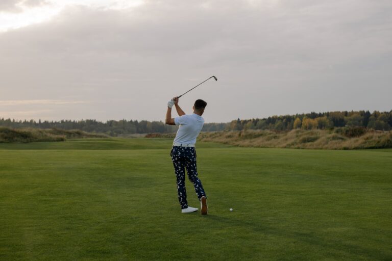 A male golfer in patterned pants swinging a club on a lush green field during sunset.
