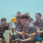 Soccer team joyfully celebrating their victory on a sunny pitch holding a trophy.