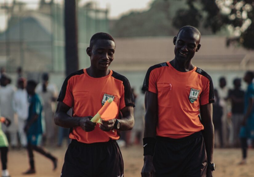 Two soccer referees in orange jerseys walking during a daytime match.