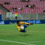 A goalkeeper in action diving to catch a soccer ball during a match in a large stadium.