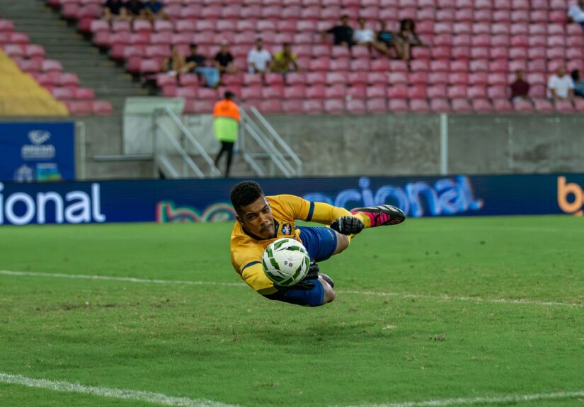 A goalkeeper in action diving to catch a soccer ball during a match in a large stadium.