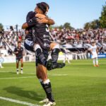 Two soccer players joyfully celebrate a goal during a match on a sunny day.