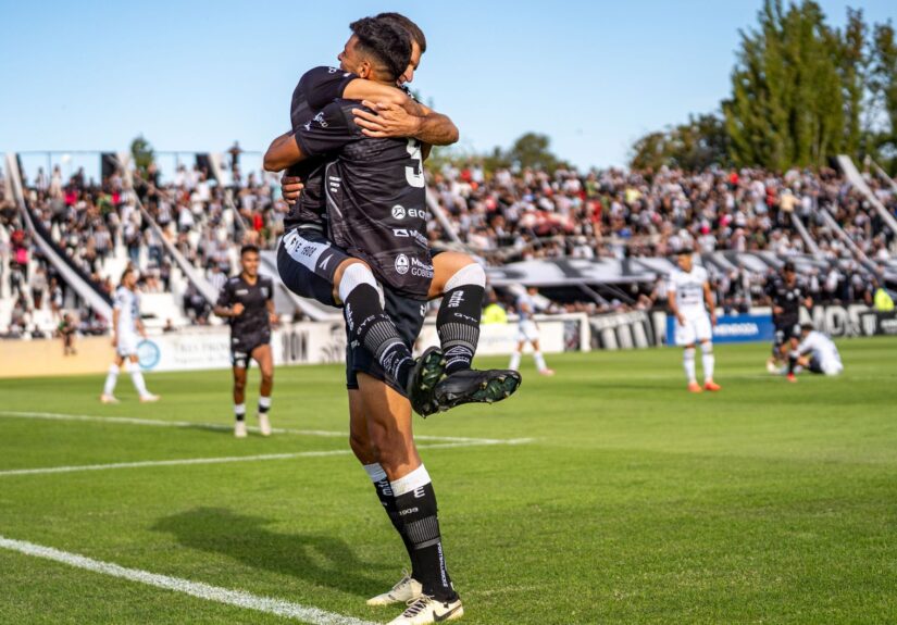 Two soccer players joyfully celebrate a goal during a match on a sunny day.