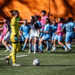 A women's soccer team celebrating after scoring a goal during a daytime match.