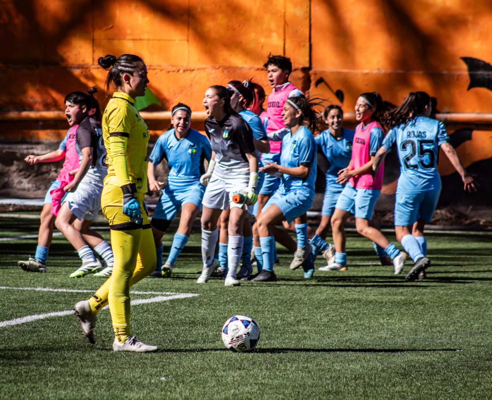 A women's soccer team celebrating after scoring a goal during a daytime match.