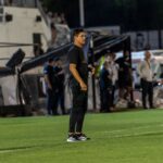 Focused football coach stands on the field sidelines during an intense match at night.