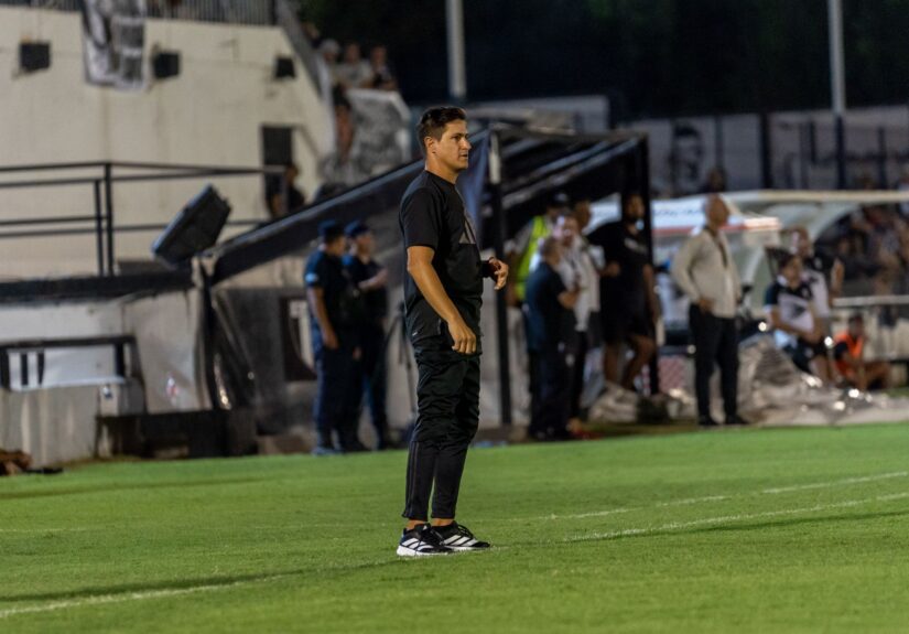 Focused football coach stands on the field sidelines during an intense match at night.