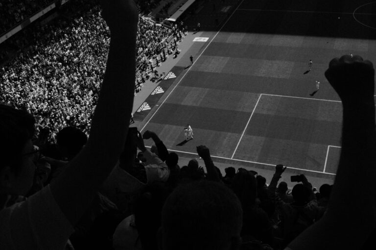 Fans cheering during a high-energy soccer match with players on the field, captured in black and white.