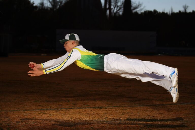 Cricketer in full sports gear leaps through the air to catch the ball, showing athleticism and focus.