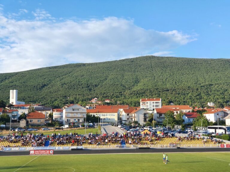 Crowded football stadium in Posušje with lush hills and townscape in the background.