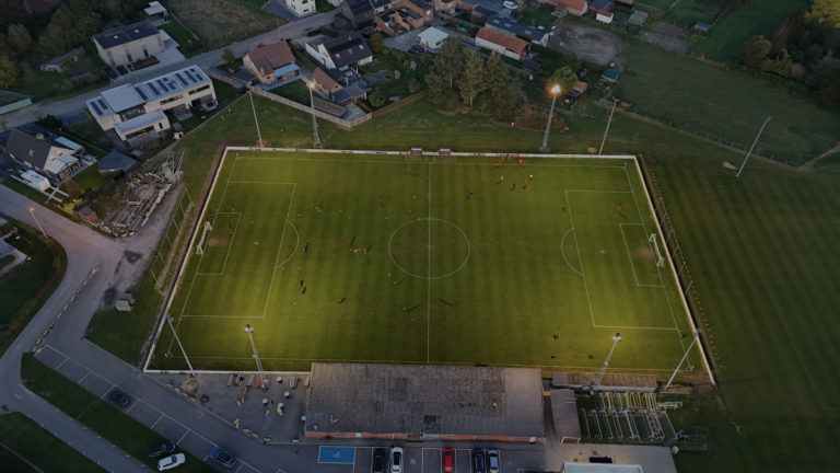Aerial shot of a soccer field at dusk in België with players practicing under floodlights.