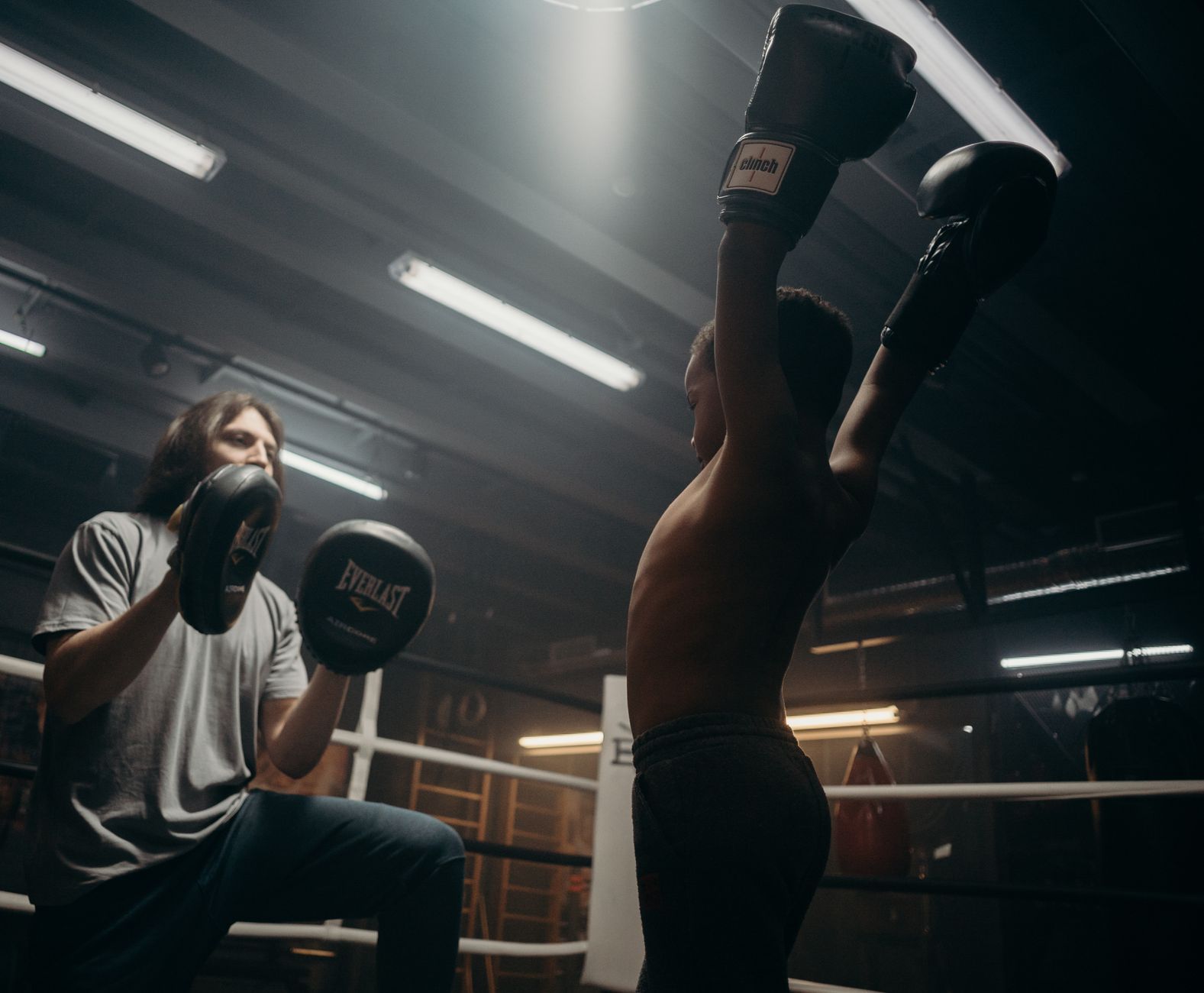 Child boxer triumphantly raises arms during boxing training session indoors.