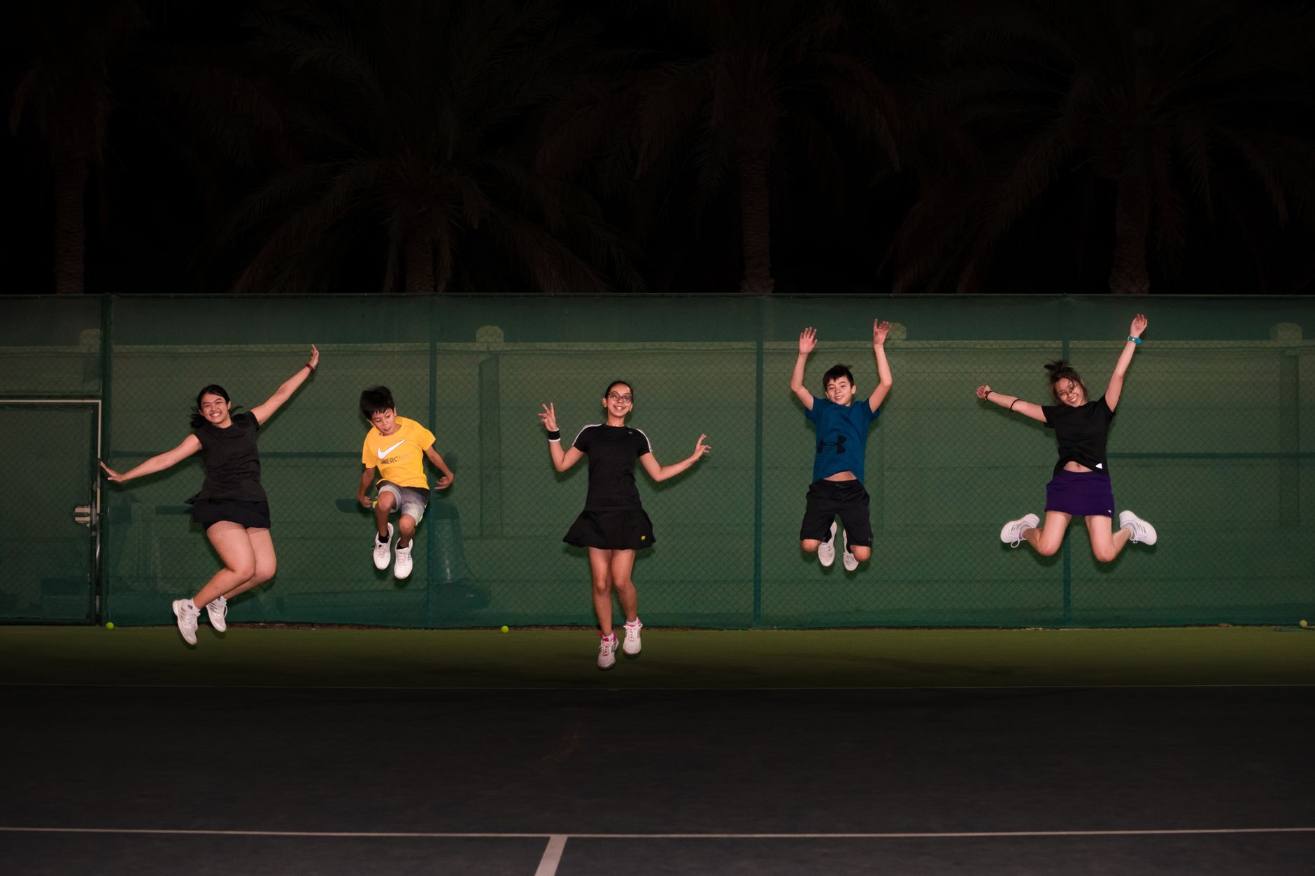 Group of teenagers jumping in celebration on an outdoor tennis court at night.