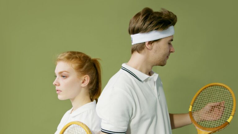 Caucasian couple in white tennis attire posing back to back with wooden rackets against a green backdrop.