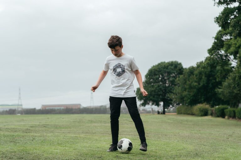 Full body of focused boy playing football in green field against trees in daytime