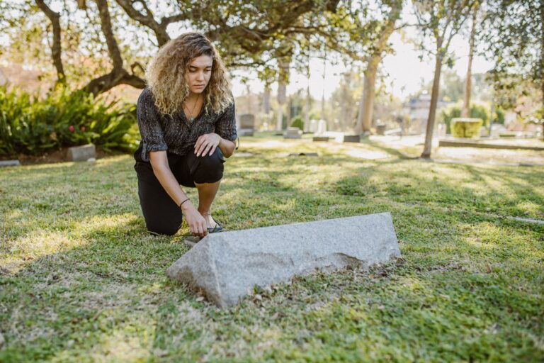 A woman kneels by a gravestone in a serene cemetery, experiencing reflection.