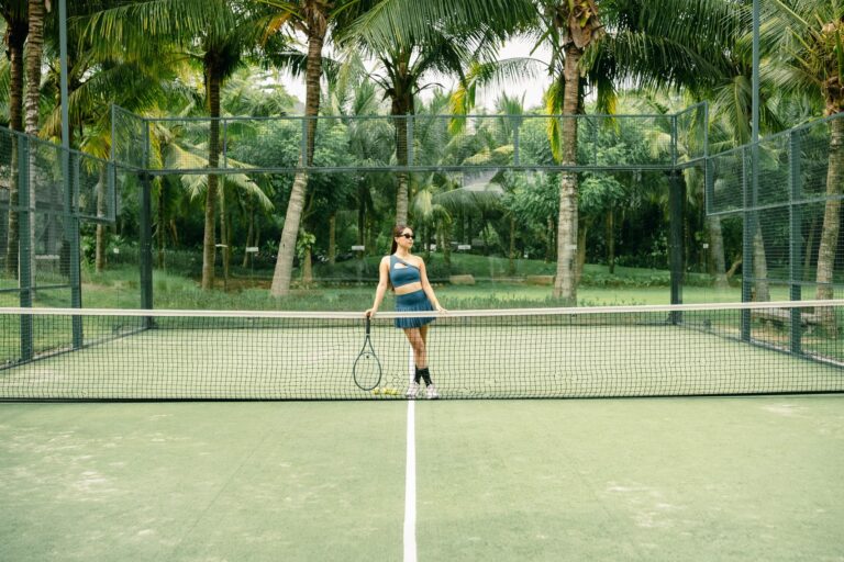A woman poses with a tennis racket on a palm tree-lined tennis court outdoors.