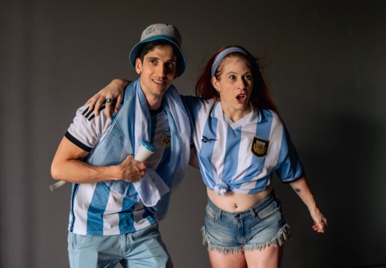 Two cheerful Argentina soccer fans celebrate wearing team jerseys indoors.