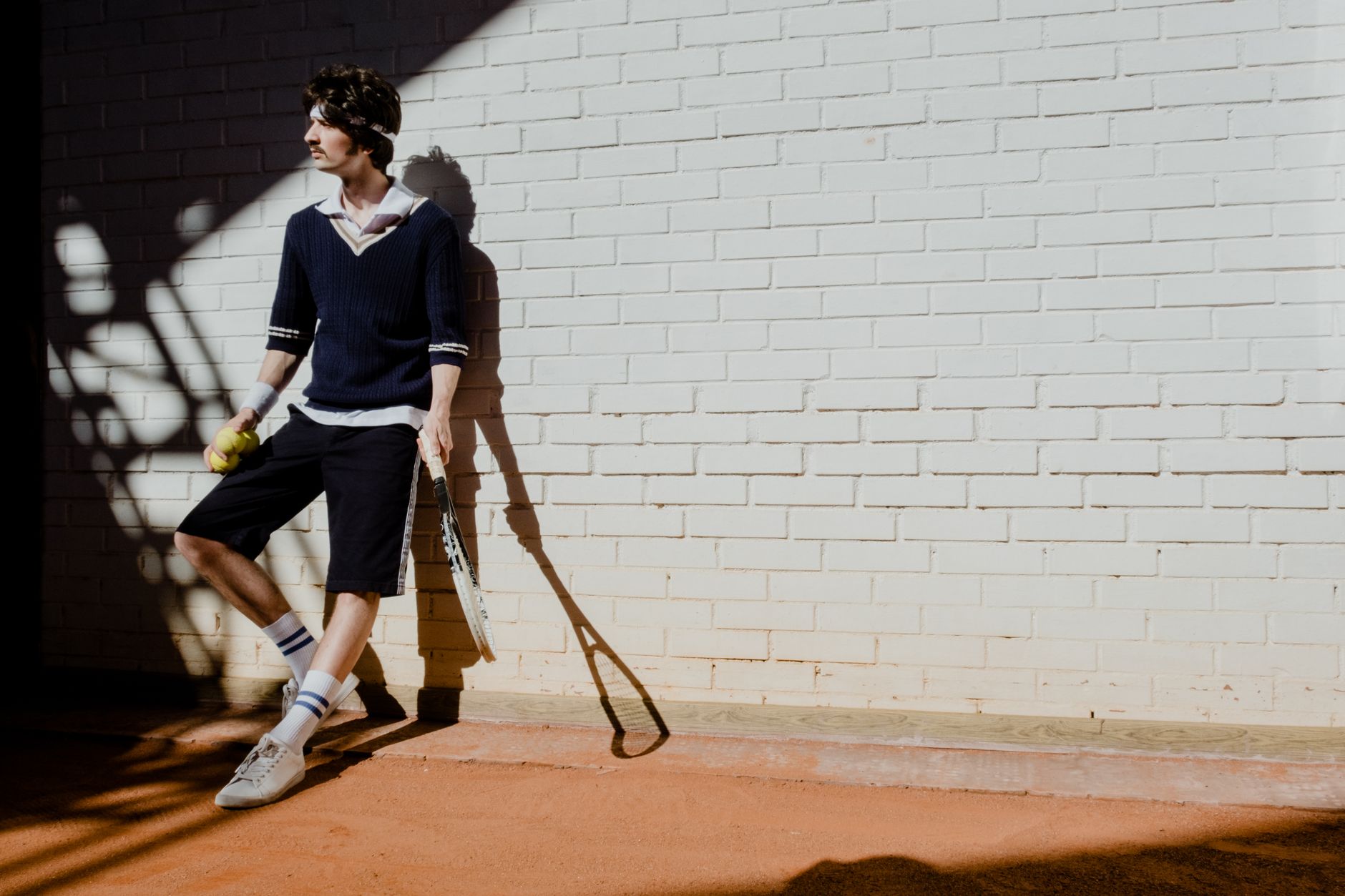 Young man in sportswear leaning against a wall holding a tennis racket and ball, bathed in sunlight.