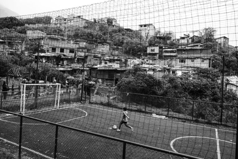 Black and white photo of a boy playing soccer on a field in Bogotá, Colombia, surrounded by urban housing.