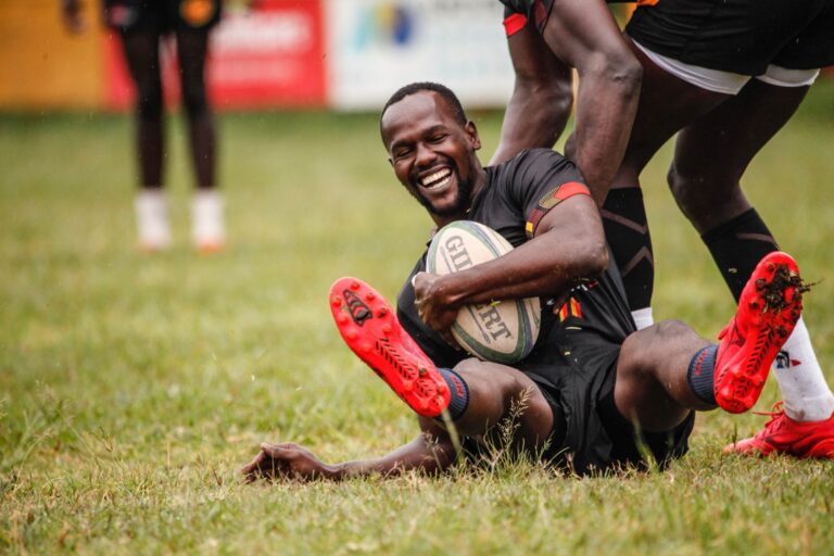 Smiling rugby player celebrating a successful try on a grassy outdoor field.
