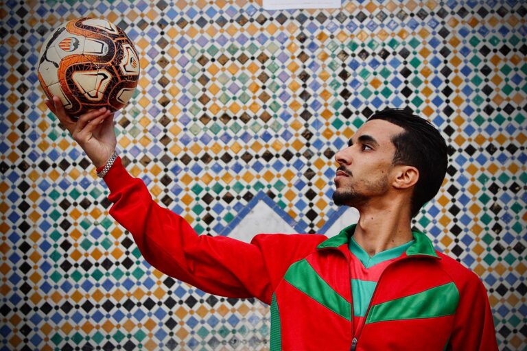 Young man in a red jacket holding a soccer ball against a colorful mosaic wall background.