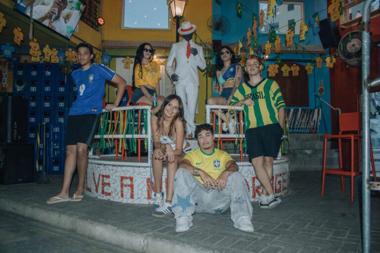 Seven young adults pose in Brazil jerseys at a vibrant, colorful outdoor venue filled with festive decorations.