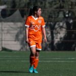 A female soccer player in orange uniform during a match on an outdoor field in Chile.