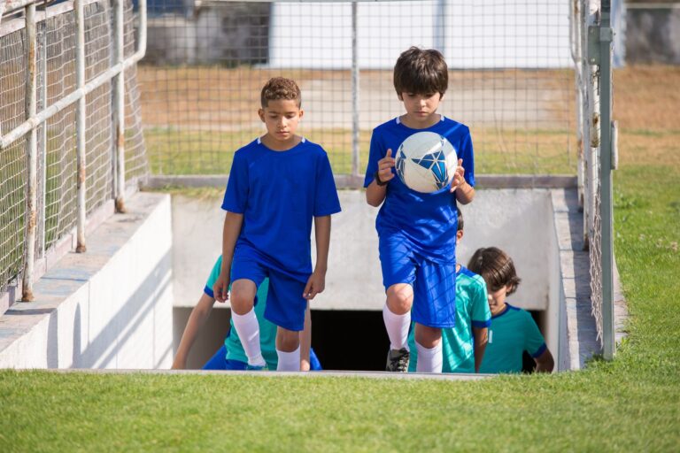 Children walking onto a soccer field during a sunny day, holding a soccer ball.