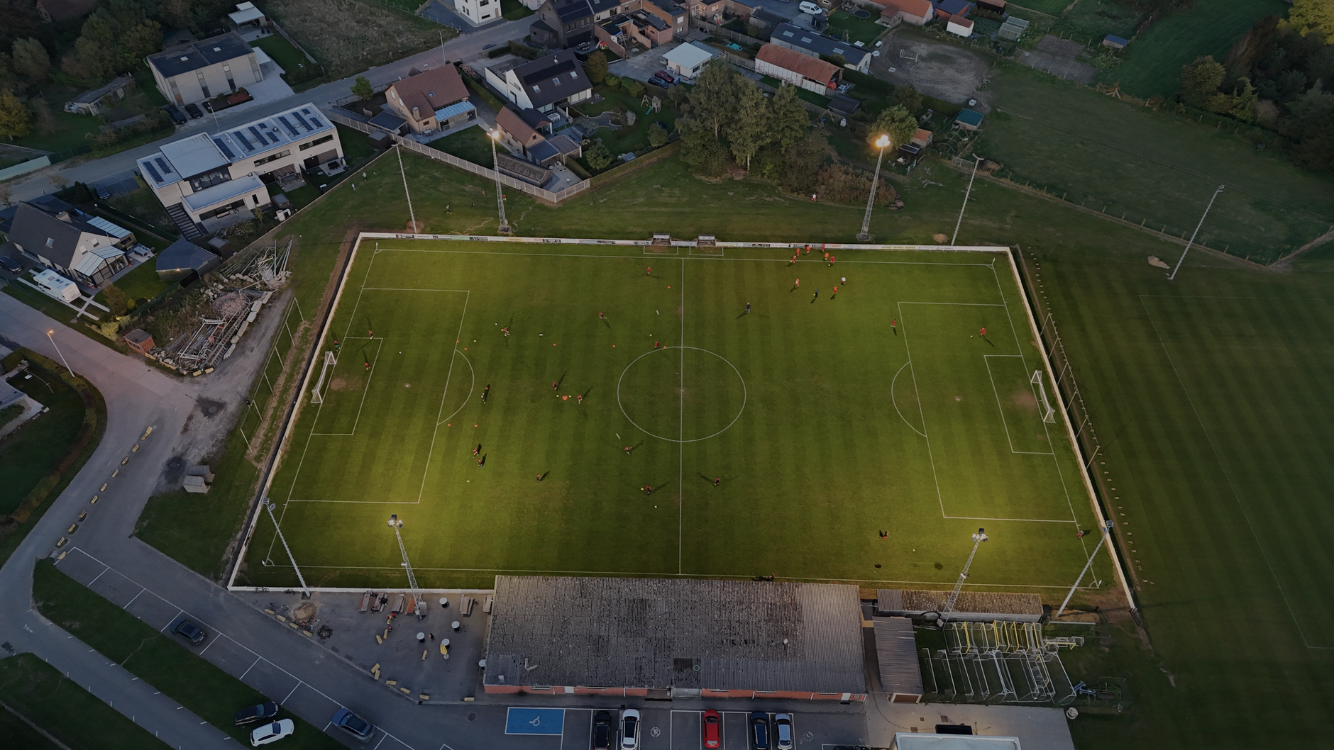 Aerial shot of a soccer field at dusk in België with players practicing under floodlights.
