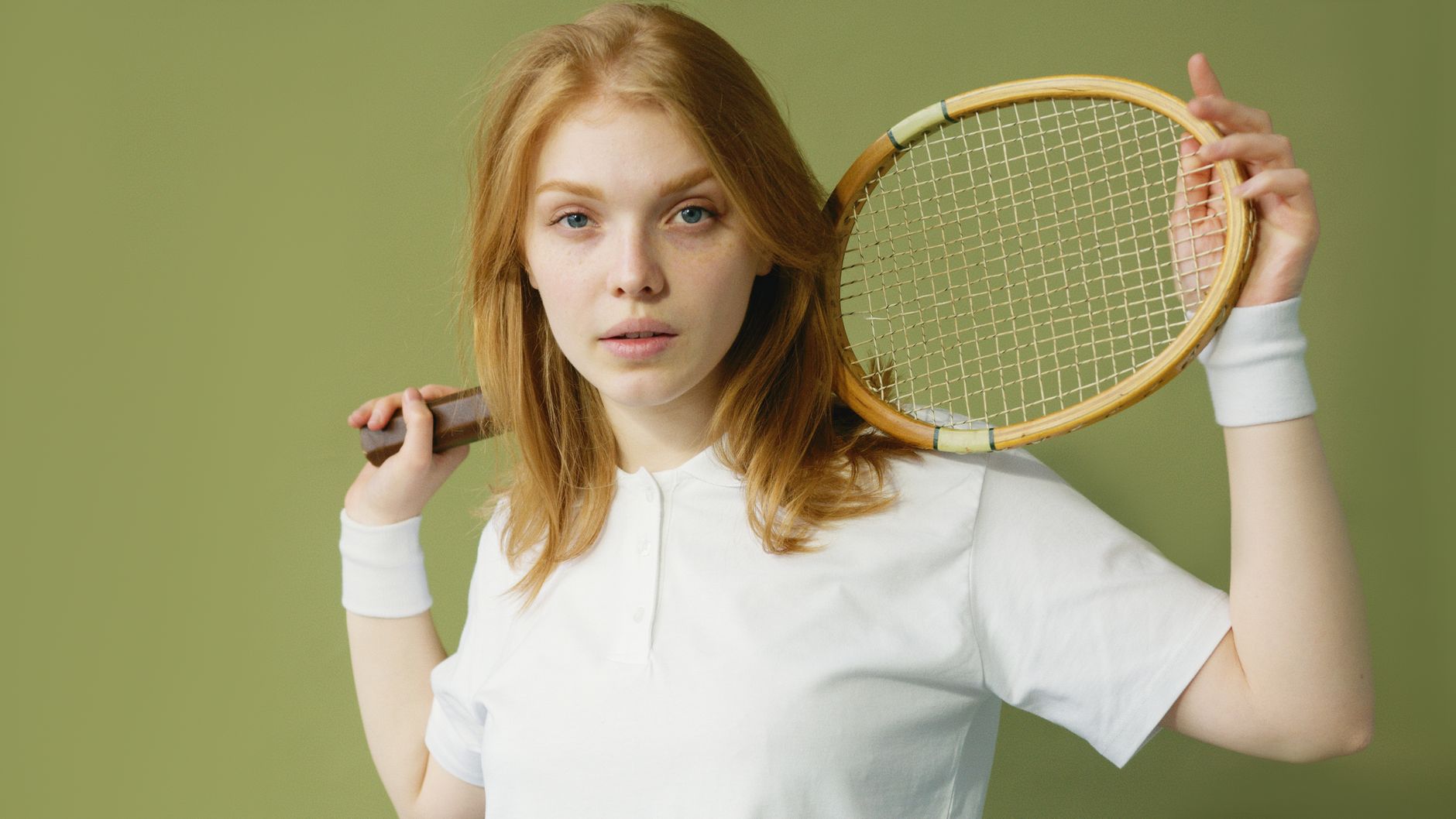 Portrait of a blonde woman with a tennis racket on her shoulder against a green background.