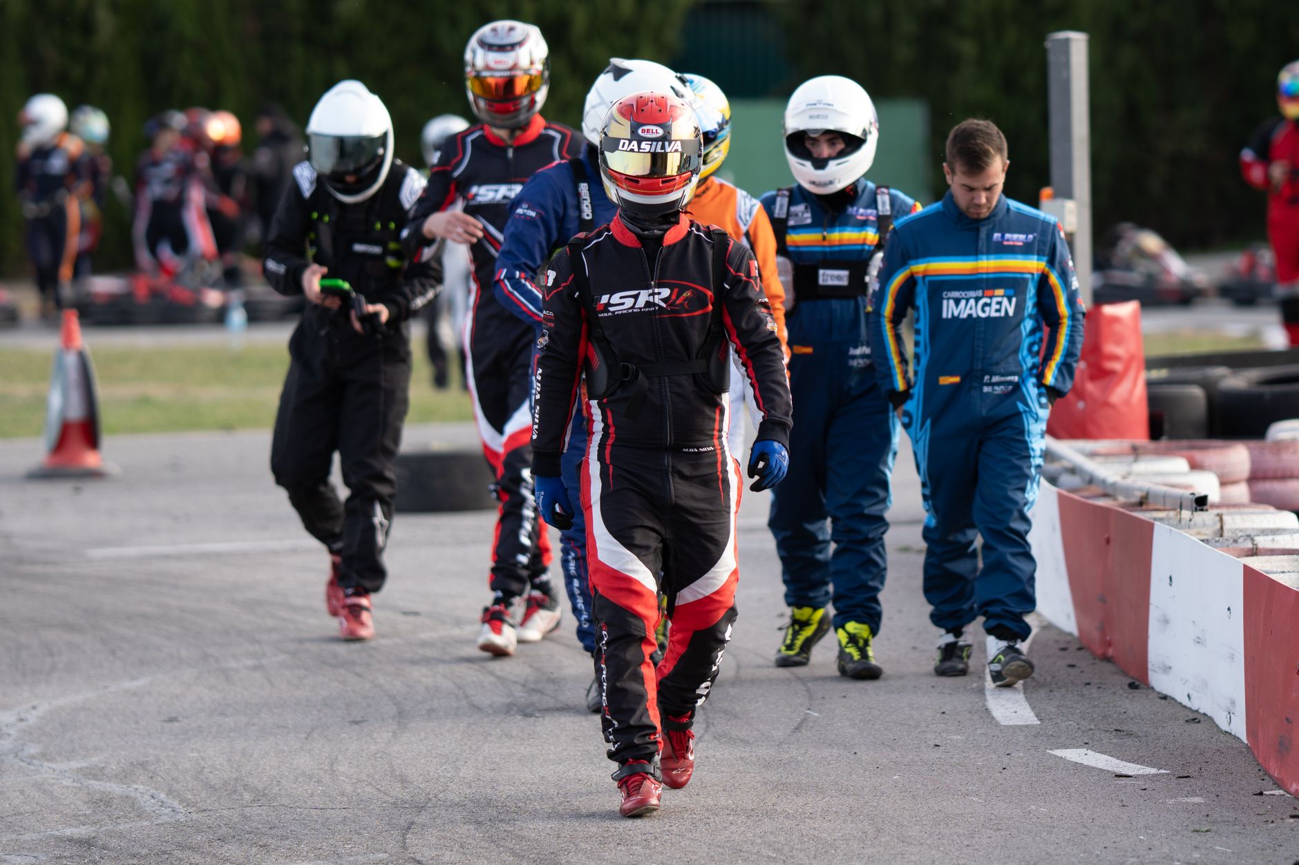 Group of go-kart racers walking on track in racing suits and helmets, signaling an exciting motorsport event.