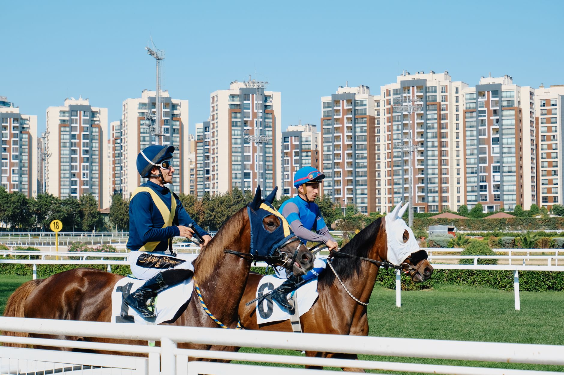 Jockeys on racehorses at a racetrack with a city skyline, combining sport and architecture.